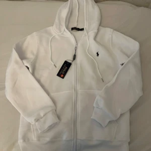 Vit hoodie från Polo Ralph Lauren - Vit hoodie från Polo Ralph Lauren med dragkedja och huva. Klassisk svart logga broderad på bröstet, snörning i huvan och ribbade muddar. Tillverkad i mjuk bomull, perfekt för chill dagar. Snygg och enkel att matcha med jeans eller joggers.