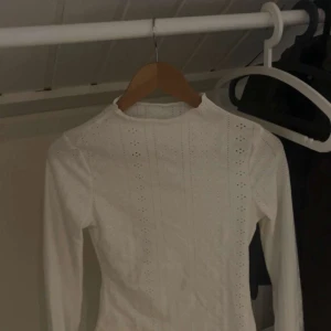 Vit långärmad blus med hålmönster - Säljer en vit långärmad blus i storlek S med snyggt hålmönster i vertikala rader. Blusen har en hög hals och är gjord i mjukt material med stretch, 95% polyester och 5% elastan. Perfekt för dig som gillar stilrena och feminina detaljer.