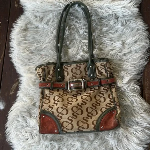 Snyggaste vintage väska❤️ - Snygg vintage handväska med brunt S-mönster, gröna skinnhandtag och detaljer, samt rödbruna mockapartier och bälte med metallspänne. Rymligt beige innerfoder med fickor och dragkedja upptill. Perfekt för dig som gillar statement-väskor med coola detaljer. Italienskt dyrt märke. Kan finnas nån fläck slitage som inte syns tydligt se bilderna❤️ Perfekt till skolan då den rymmer så mycket❤️Köpt sacond hand. Tryck gärna köp nu