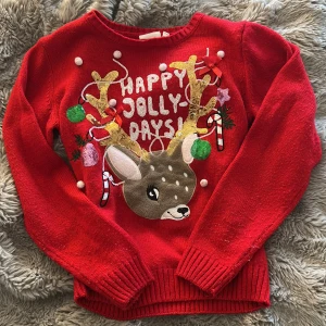 Röd jultröja med ren från KappAhl - Supermysig röd stickad jultröja från KappAhl med ett gulligt renmotiv, glittriga paljettdetaljer, rosetter och texten 'Happy Jolly Days' framtill. Tröjan har långa ärmar och ribbade muddar. Perfekt för att maxa julstämningen!