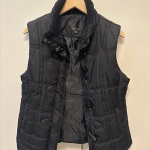 Svart quiltad dunväst från Amisu - Svart dunväst från Amisu med quiltad design och hög ribbad krage. Västen har praktiska fickor och stängs med både dragkedja och knappar. Perfekt att slänga över en hoodie för extra värme när det är kallt ute.