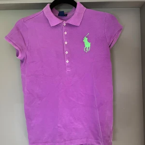 Lila pikétopp från Ralph Lauren - Säljer en lila pikétopp från Ralph Lauren med korta ärmar och klassisk krage. Toppens framsida har knappar och ett stort grönt broderat Polo-logomärke. Perfekt för dig som gillar sportig och stilren vibe.