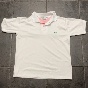Vit pikétröja från Lacoste - Vit pikétröja från Lacoste med klassisk krage och korta ärmar. Tröjan har den ikoniska gröna krokodilloggan broderad på bröstet och en snygg rosa detalj på insidan av kragen. Perfekt för en clean och sportig look.