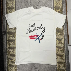 Saint Laurent ”No Smoking” T shirt - Vit t-shirt från Saint Laurent med coolt grafiskt tryck framtill. Trycket består av svarta bokstäver, röda läppar och blå detaljer. T-shirten har rund hals och korta ärmar, tillverkad i mjuk bomull. Perfekt för dig som gillar statement-plagg. Storlek M, aldrig använd.
