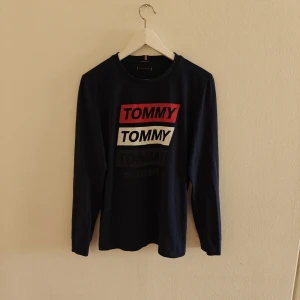 Tommy Hilfiger tröja - Snygg mörkblå långärmad tröja från Tommy Hilfiger med tryck i rött, vitt och svart på bröstet. Tillverkad i 100% ekologisk bomull, rund hals och klassisk passform. Perfekt för dig som gillar stilrena logotröjor med street-vibe.