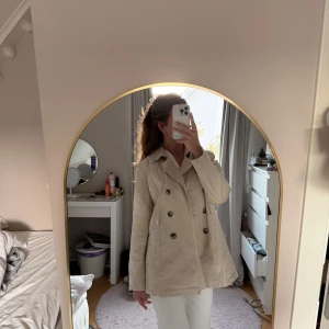 Beige trenchcoat med knappar - Snygg beige trenchcoat med dubbelknäppning och klassisk krage. Jackan har två sidofickor och stora bruna knappar framtill. Materialet är mjukt och känns som bomull, perfekt för dig som gillar stilrena och tidlösa plagg. Passar dig som vill ha en chill och clean look.