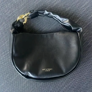 Svart axelväska från Art Street London - Snygg svart axelväska i skinnimitation med mjuk, rundad form och bred axelrem med chunky guldfärgad kedjedetalj. Väskan har dragkedja upptill och en extra ficka med dragkedja på baksidan. Perfekt storlek för det viktigaste och stilren look.