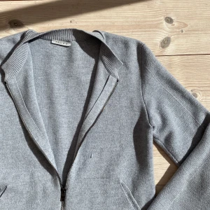 Grå kofta med dragkedja från Falke - Snygg grå kofta från Falke med hel dragkedja framtill. Mjuk stickad känsla och långärmad modell som passar perfekt för kyliga dagar. Enkel och stilren design med ribbade muddar vid ärmslut och nederkant. Perfekt att slänga på sig när du vill vara både chill och stilren. Har några defekter men syns på bilderna