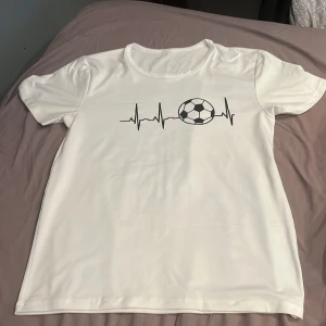 Vit t-shirt med fotbollstryck - Vit t-shirt med rund hals och korta ärmar. På framsidan finns ett coolt tryck med hjärtslag och en fotboll, perfekt för dig som älskar sporten. Materialet känns mjukt och lätt, troligen polyester eller bomull. Enkel och sportig stil som passar till träning eller häng.