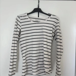 Randig långärmad tröja Basic You - Vit långärmad tröja från Basic You med svarta horisontella ränder. Tillverkad i mjuk ekologisk bomull, har en rund halsringning och normal passform. Perfekt för dig som gillar enkel och stilren look med lite fransk vibe.