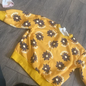 Gul sweatshirt med blommor från SHEIN - Cool gul sweatshirt från SHEIN med stora mörkbruna blommor och texten 'SUNSHINE' tryckt över hela tröjan. Ribbad mudd vid hals, ärmslut och nederkant. Perfekt för dig som gillar färg och mönster!