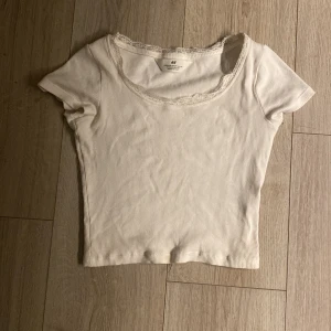 Vit ribbad topp med spets från H&M - Säljer en söt vit ribbad topp från H&M i storlek 134/140. Toppen har korta ärmar och en rundad halsringning med spetsdetalj som ger en extra touch. Perfekt att styla med jeans eller kjol för en chill look.