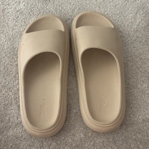 Beige sandaler från Zara, strl 37 - Säljer ett par chunky beige sandaler från Zara i storlek 37. De har öppen tå, platt sula och bred rem över foten. Tillverkade i syntetmaterial med en clean och modern look, perfekta för varma dagar. Sulan har tydliga ränder och Zara-logga undertill.