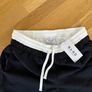 Svarta kostymbyxor från NA-KD - Svarta kostymbyxor från NA-KD med vit, ribbad midja och snörning. Byxorna har raka ben, sidofickor och en bakficka. Perfekta för chill dagar och har en clean, stilren look med kontrast i midjan. Materialet är mjukt och skönt, troligtvis bomull eller polyester.