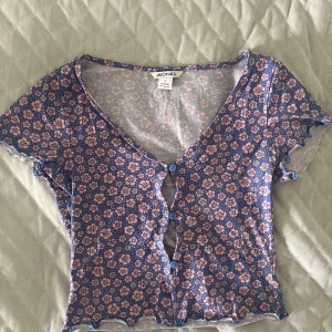 Blommig topp från Monki - Supersöt topp från Monki i blått med rosa blommigt mönster. V-ringad med korta ärmar och blå knappar framtill. Materialet är mjuk bomull med lite stretch för skön passform. Perfekt för dig som gillar färg och mönster!💙🩷Axelbredd: 36 cm Längd: 42 cm