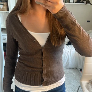 Brun kofta från H&M med v-ringning - Snygg brun kofta från H&M med v-ringad hals och knappar framtill. Modellen är långärmad och har ribbade muddar vid ärmslut och nederkant. Perfekt att slänga över en t-shirt eller linne för en chill look. Materialet är mjuk bomull och polyester. Står att storleken är 36, men skulle säga 34/xs. Skriv vid frågor!🥰
