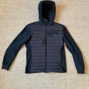 Svart dunjacka med huva från UBR - Snygg svart dunjacka från UBR i storlek M. Jackan har quiltad framsida, hel dragkedja och praktisk huva. Materialet är lätt och smidigt, perfekt för kyliga dagar. UBR-logga på bröstet och två sidofickor med dragkedja. Stilren och sportig look.