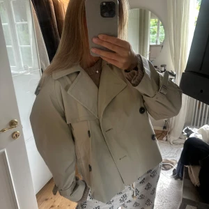 Trench - Snygg kort trenchcoat i beige med bred krage, dubbelknäppning och stora svarta knappar. Jackan har bälte vid ärmsluten och en loose passform som ger en chill vibe. Perfekt för dig som gillar klassisk stil med modern twist.