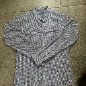 Rutig skjorta från Polo Ralph Lauren - Snygg rutig skjorta från Polo Ralph Lauren i klassisk svartvit färg med rött broderat logomärke på bröstet. Skjortan har lång ärm, knappar framtill och krage. Perfekt för dig som gillar stilrena och tidlösa plagg.