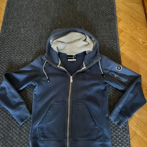 Marinblå hoodie från Pelle P - Snygg marinblå hoodie från Pelle P med dragkedja, huva med grått foder och coolt emblem på ärmen. Tröjan har praktiska fickor fram och snörning i huvan. Perfekt för chill dagar och när du vill ha en sportig look.