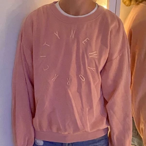 Rosa sweatshirt med New York-tryck - Säljer en rosa sweatshirt med rund hals och trycket 'New York City' i cirkel på bröstet. Tröjan har långa ärmar och ribbade muddar vid ärmslut och nederkant. Perfekt för chill dagar och enkel att matcha med jeans.