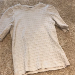 Vit ribbad topp från Name It - Säljer en vit topp från Name It i storlek 140. Den har ett ribbat, strukturerat tyg och puffade axlar som ger en snygg touch. Ärmarna är trekvarts och modellen är normal i passningen. Perfekt för dig som gillar stilrena och enkla plagg.