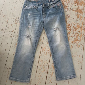 Miss Me ljusblå bootcut jeans med broderi - Säljer ett par ljusblå jeans från Miss Me med bootcut passform med lite kortare benlängd och slitna detaljer framtill. Baksidan har snygga broderade fickor med mönster och dekorativa knappar. Jeansen har låg midja och är tillverkade i klassiskt denimtyg. Perfekt för dig som gillar unik stil!