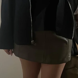 Olivgrön skinnkjol i kort modell - Snygg olivgrön minikjol i skinnimitation med raka sömmar och bälteshällor. Bra skick! 