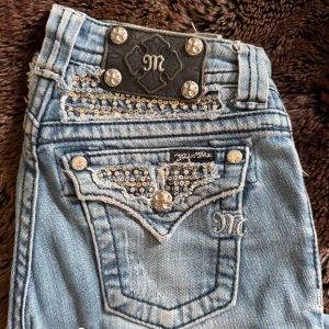 Miss Me ljusblå lowaist bootcut jeans med paljetter - Säljer ett par ljusblå jeans från Miss Me med bootcut passform. Jeansen har snygga paljett- och nitdetaljer på fickorna, samt dekorativa knappar och broderad logga. Materialet är klassisk jeans med stretch och slitningar. Lågmidjade bootcut jeanse passar xs! 