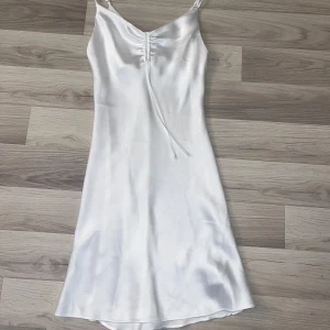 Vit satin klänning från Bik Bok XS - Supersnygg vit klänning från Bik Bok i satin med smala axelband och snörning framtill. Klänningen har en slank siluett och är lätt utsvängd nertill, perfekt för dig som gillar att klä upp dig utan att det blir för mycket! Den har endast använts 1 gång.