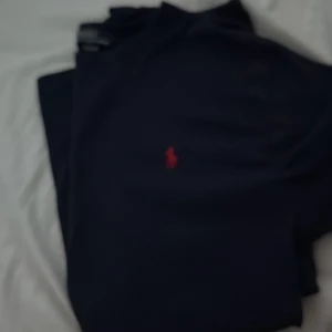 Blå T-shirt  från Ralph Lauren - Klassisk svart pikétröja från Ralph Lauren med den ikoniska röda logon broderad på bröstet. Perfekt för dig som gillar stilrena och tidlösa plagg. Tröjan har korta ärmar och en bekväm passform som funkar till många olika looks. Barnstorlek passar xs/s