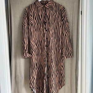 Zebramönstrad långklänning i beige och svart - Snygg långklänning med zebramönster i beige och svart. Klänningen har långa ärmar, knappar framtill och en avslappnad passform. Perfekt för dig som vill sticka ut med ett djurmönster och cool stil.