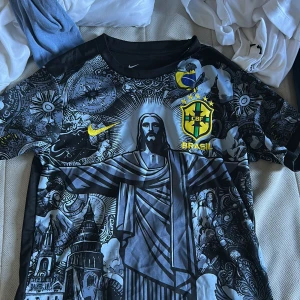Nike Brasil CBF Dri-Fit fotbollströja - Unik Brasil CBF fotbollströja från Nike med Dri-Fit material. Svart och grå bas med detaljerat grafiskt tryck av Kristusstatyn och brasilianska motiv, samt gula loggor och emblem. Kortärmad och perfekt för dig som vill sticka ut på planen.