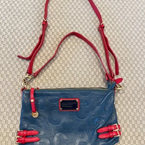 Blå/röd väska från Marc Jacobs - Cool blå väska med röda axelband och detaljer från Marc Jacobs. Mässingsspännen och dubbla axelband i olika längder.  1 mittenfack med magnetknapp. 2 stora fack med dragkedja och ett mindre fack med dragkedja på baksidan. Mått: 30x19 cm. Liten vit fläck på insidan av mittenfacket, se bild.