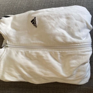Vit träningsjacka från Adidas - Säljer en vit träningsjacka från Adidas med svarta ränder längs ärmarna och dragkedja framtill. Perfekt för sportiga aktiviteter eller en avslappnad stil. Passar som både M och S, priset kan diskuteras.