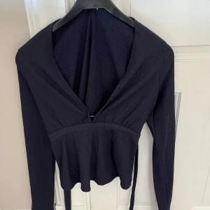 Svart ribbad topp - Snygg svart ribbad topp med långa ärmar och v-ringad design. Perfekt för en stilren look. Toppen har en knytning i midjan som ger en fin passform.