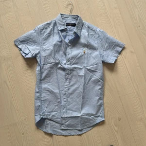 Kortärmad Blå Ralph Lauren Skjorta - Säljer denna kortärmade blåa Ralph lauren skjorta då den inte längre kommer till användning. Inga skador så i princip nyskick. Perfekt nu inför sommaren. Skriv gärna vid fler frågor:)