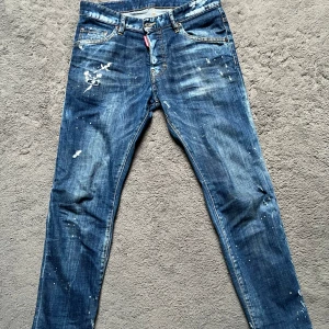 Blå jeans från Dsquared2 - Snygga blå jeans från Dsquared2 med unika slitningar och färgstänk. De har en klassisk femficksdesign och en röd logotypdetalj vid gylfen. Perfekta för en trendig look.