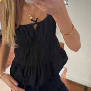 Svart ärmlös topp med volanger - Snygg svart ärmlös topp med volanger och knytning fram. Perfekt för en stilren look. Passar bra till både jeans och kjol.