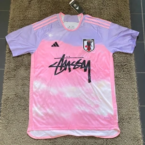 Stussy X Adidas Japan T-shirt - Säljer en riktigt fet fotbollströja från Adidas i rosa och lila med Japan JFA-logga och Stüssy-tryck. Tröjan är kortärmad och har en modern design med Aeroready-teknologi för bästa komfort. Riktigt fräsch till sommaren.
