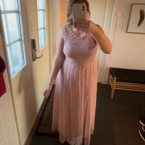 Ljusrosa långklänning med spetsdetaljer - Elegant ljusrosa långklänning med spetsdetaljer på överdelen. Klänningen är ärmlös och har en flödande kjol som ger en romantisk känsla. Perfekt för bal och sommarens festligheter! Klänningen är för liten för mig, därför sitter den lite tajt. Har man storlek 38 så sitter den inte lika tajt.
