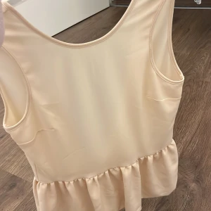 Beige ärmlös topp med volang - Snygg beige ärmlös topp med djup v-ringning och volangdetalj i nederkant. Perfekt för en stilren look.