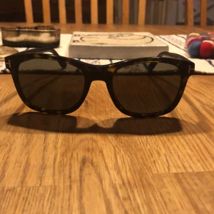 Solglasögon från Tom Ford - Snygga solglasögon från Tom Ford med en klassisk design. Bågarna är i en mörk färg med en subtil sköldpaddsmönster och har en elegant T-detalj vid tinningarna. Perfekta för en stilren look.