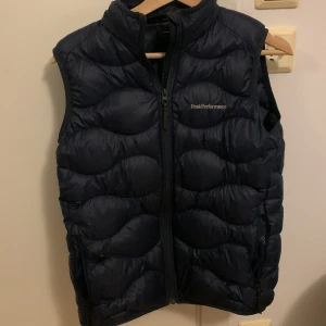 Svart dunväst från Peak Performance - Snygg svart dunväst från Peak Performance med dragkedja framtill. Perfekt för kyligare dagar och ger en sportig look. Västen har en quiltad design som ger extra värme och stil. Den har en liten öppning som vi har tejpat över men du får göra som du vill annars i bra skick 