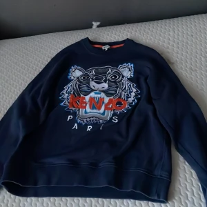 Mörkblå sweatshirt från Kenzo - Säljer en mörkblå sweatshirt från Kenzo med ett stort tigerbroderi och logotyp på framsidan. Tröjan har en normal passform och är tillverkad i ett mjukt material. Perfekt för en stilren och avslappnad look.tröjan är knappt använd har haft på mig den kanske fem gånger.det är small i barn storlek.