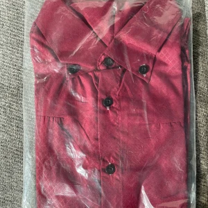 Vinröd skjorta med knappar - Snygg vinröd skjorta med svarta knappar. Skjortan har en klassisk krage och är förpackad i plast, helt oanvänd.