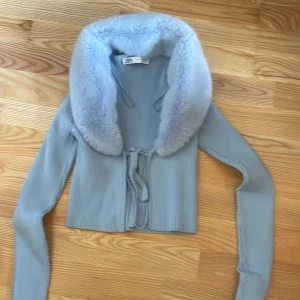 Ljusblå ribbad kofta med fuskpäls - Säljer en ljusblå ribbad kofta med en lyxig fuskpälsdetalj runt kragen. Koftan är croppad och har långa ärmar samt knytband framtill. Perfekt för att ge en stilren touch till din outfit.