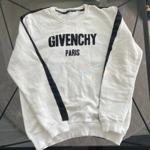 Vit sweatshirt från Givenchy - Säljer en stilren vit sweatshirt från Givenchy med svart text 'GIVENCHY PARIS' på framsidan. Tröjan har svarta detaljer längs ärmarna och ribbade muddar vid ärmslut och nederkant. Perfekt för en avslappnad och trendig look. Har tecken på använding och den är tillverkad i Turkiet.