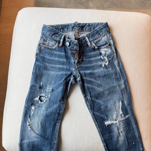 Blå jeans från DSQUARED2 - Snygga blå jeans från DSQUARED2 med slitna detaljer och färgstänk. Köpa från Farfetch och säljs nu pga för litet för mig. Strl 34. Har spruckit baktill, därav defekt, finns bild. Går lätt att ordna genom att sy dit en lapp. Har tyvärr inte hunnit göra det själv. (I övrigt är jeansen i ”bra skick”).