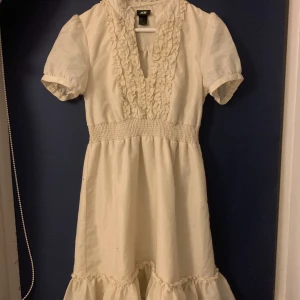 Crémevit klänning från H&M - Charmig crémevit klänning från H&M med volangdetaljer och korta ärmar. Klänningen har en v-ringad design med smock i midjan för en bekväm passform. Perfekt för sommarens alla tillfällen.